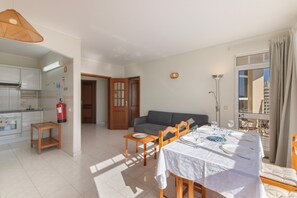 A well-lit living area with a dining table set for two, a sofa, and a kitchen in the background. - Apartment 'Camões 12 Frente Mar' with Shared Pool and Balcony (Armação de Pêra)