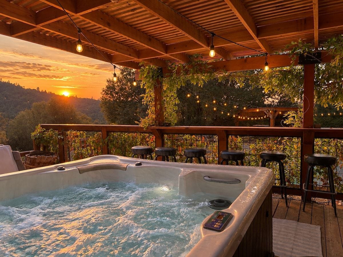 A hot tub under a wooden pergola with string lights, surrounded by bar stools and a view of the sunset.