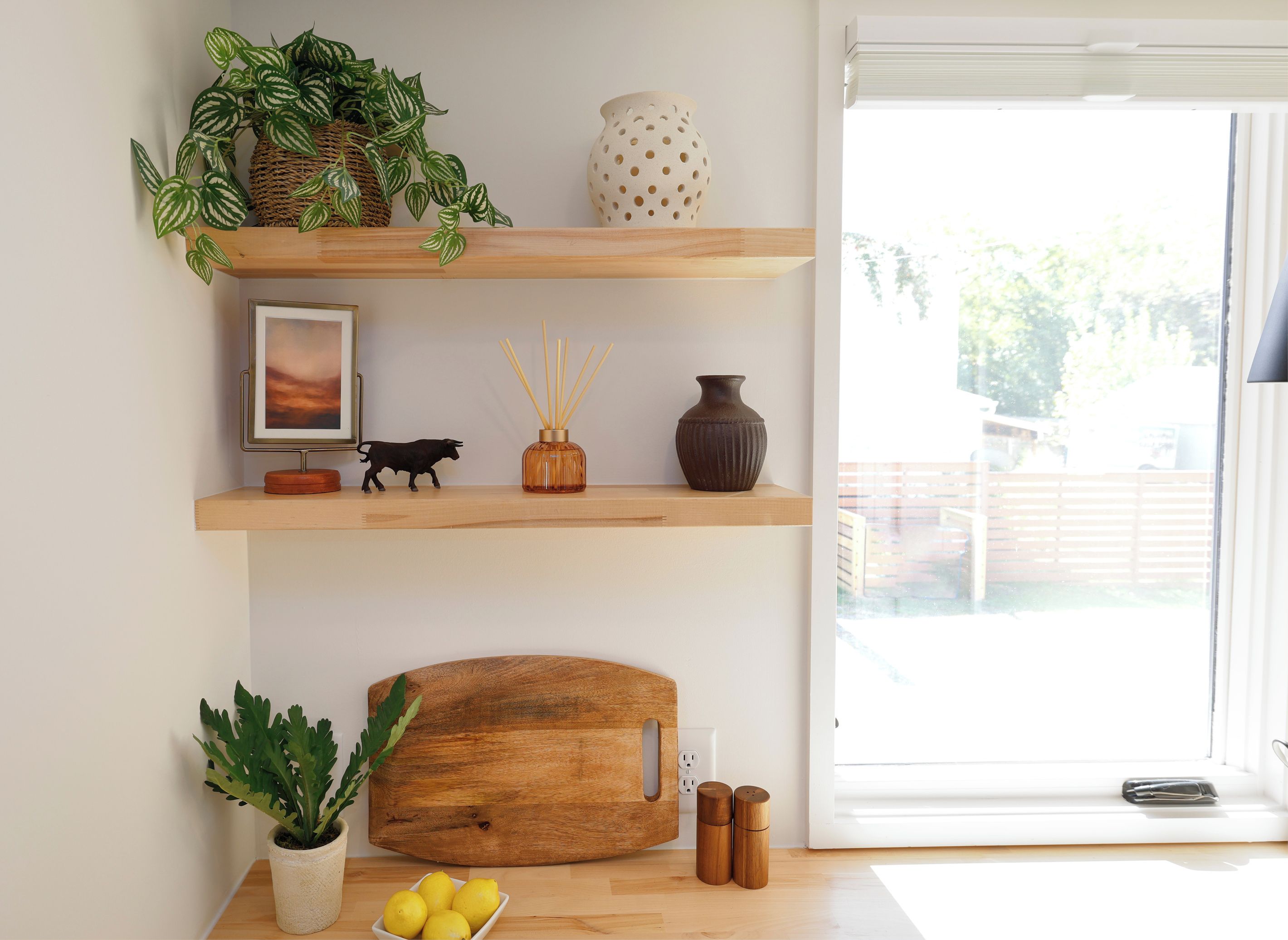 Une pièce avec une étagère en bois où sont posées des plantes, un vase, un cadre et un objet décoratif. On y trouve également une planche à découper en bois et une petite table sur laquelle se trouvent une plante en pot et des citrons.