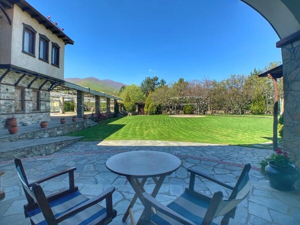 A patio with stone flooring, two chairs, a table, and a view of a well-maintained lawn and trees. - Hotel Apsis (Emporio)