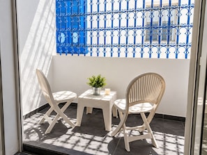 Ein Balkon mit zwei weißen Korbstühlen, einem kleinen Tisch mit einem Topfpflanzen und einem blauen Metallzaun im Hintergrund.