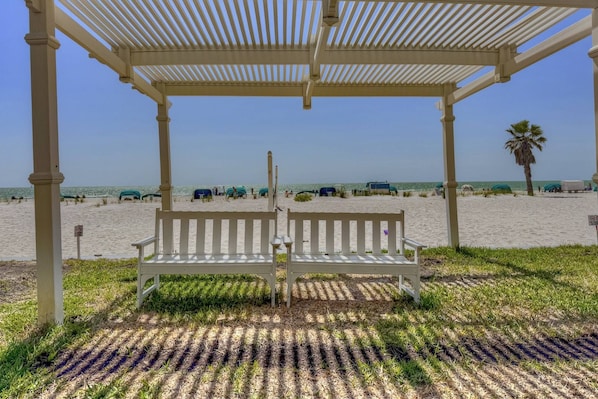 A beach gazebo with a bench, a clear view of the beach, and a palm tree in the background. - Large 6th Floor Gulf-Front Penthouse | Heated Pool | 3BR, 5 Beds, Tiki Bar & Incredible Beach Views (Treasure Island)