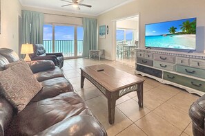 A living room with a leather sofa, a wooden coffee table, a television, and a balcony with a view of the beach. - Large 6th Floor Gulf-Front Penthouse | Heated Pool | 3BR, 5 Beds, Tiki Bar & Incredible Beach Views (Treasure Island)
