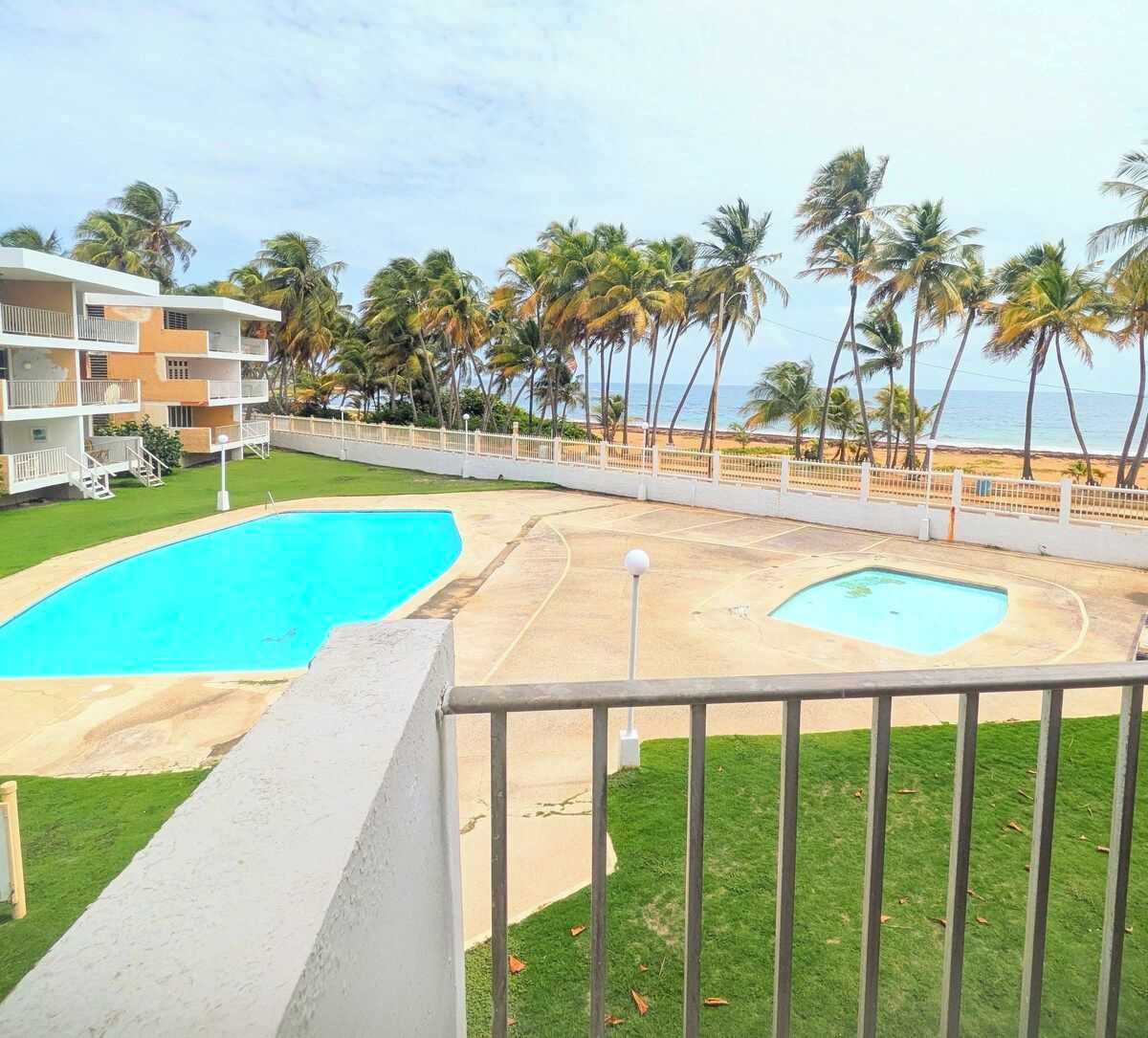 Un área de piscina con dos piscinas, un césped y vistas a la playa con palmeras.