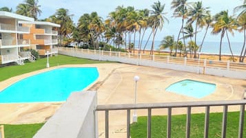 Un área de piscina con dos piscinas, un césped y vistas a la playa con palmeras.
