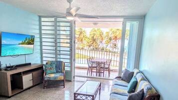 Una sala de estar con televisor, un sofá, un sillón, una mesita de centro y un balcón con vistas a palmeras.