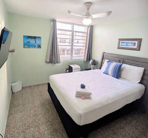 Una habitación de hotel con cama, ventilador de techo, ventana con cortinas, televisor montado en la pared y un pequeño cubo de basura.