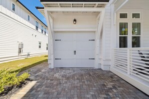 A white garage door with a black handle, a small outdoor light fixture above it, and a brick-paved driveway leading to the garage. - The Sandcastle at Bluewater Landing (Santa Rosa Beach)