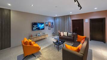 A modern living room with a flat-screen TV, two sofas with orange cushions, a coffee table, and a large window with curtains.