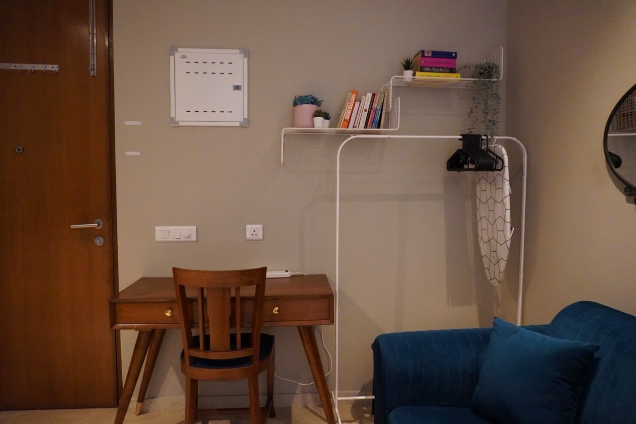 A room with a wooden desk, a blue sofa, a white shelf with books and plants, and a wall-mounted light fixture.