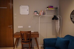 Een kamer met een houten bureau, een blauwe bank, een wit schap met boeken en planten, en een wandlamp.