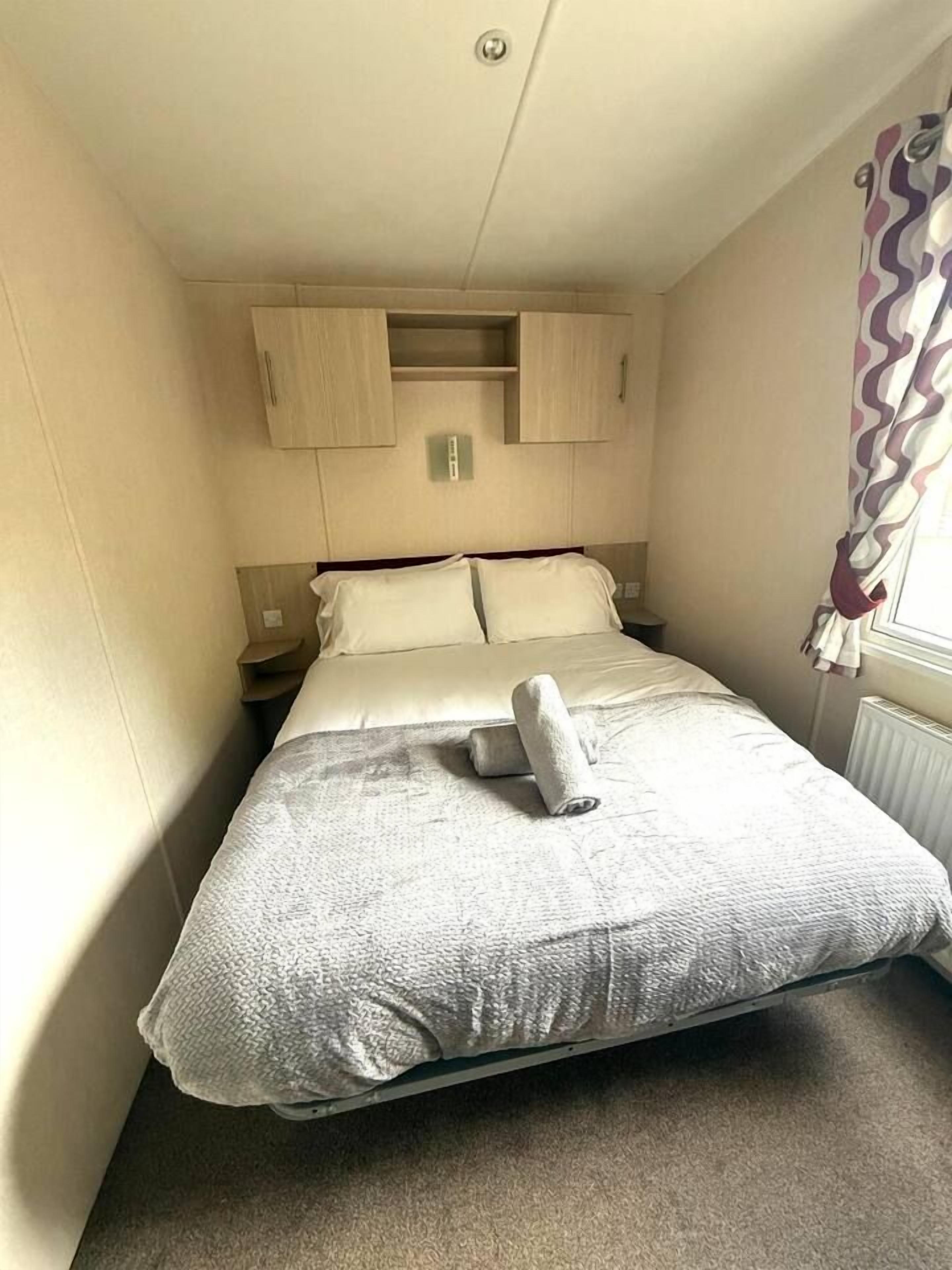 Un lit simple avec une couette blanche et une serviette enroulée, une tête de lit en bois, une table de chevet et une fenêtre avec des rideaux.
