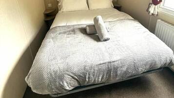 Un lit simple avec une couette blanche et une serviette enroulée, une tête de lit en bois, une table de chevet et une fenêtre avec des rideaux.