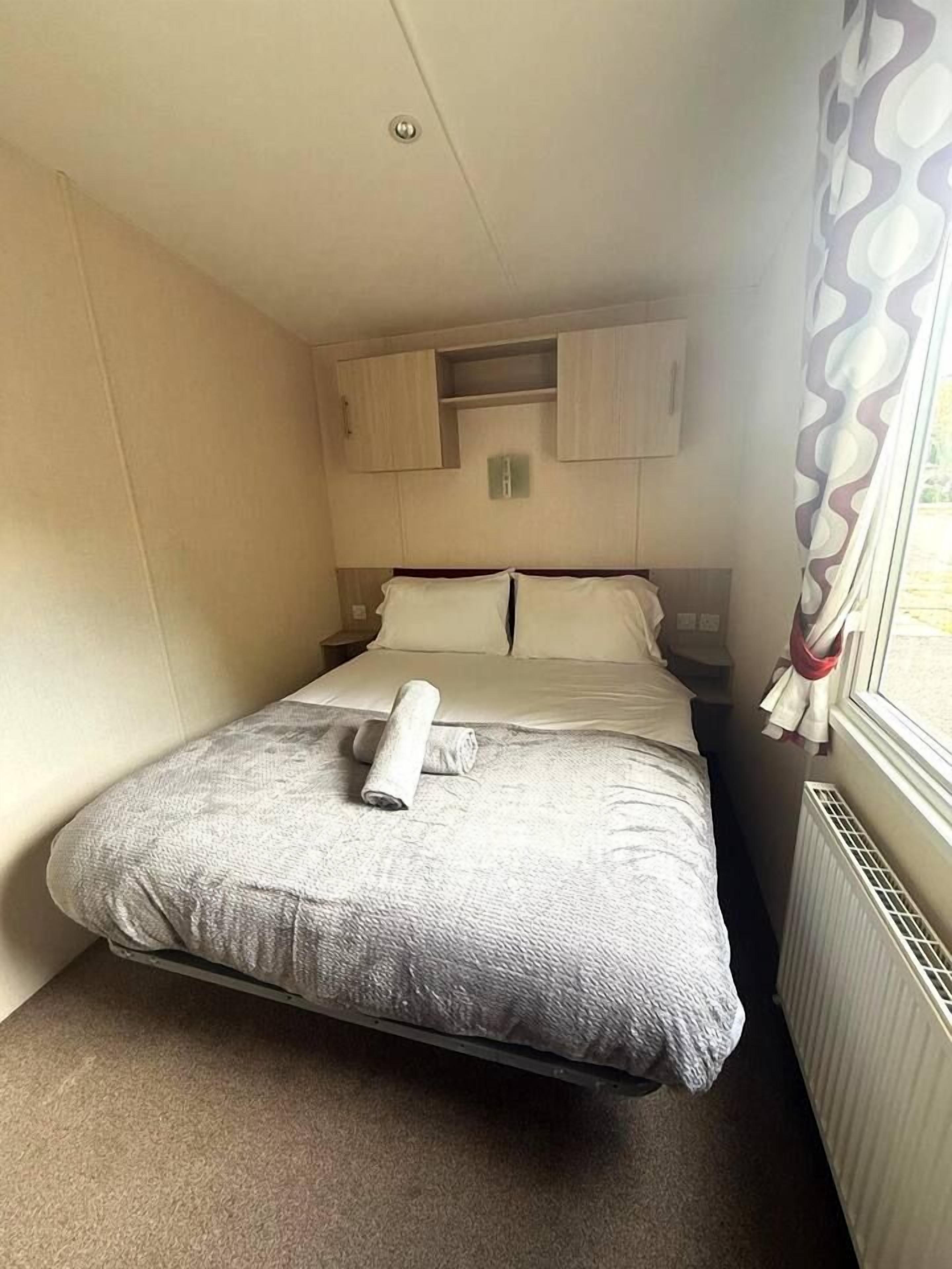 A single bed with a rolled towel, a nightstand, a window with curtains, and a radiator.