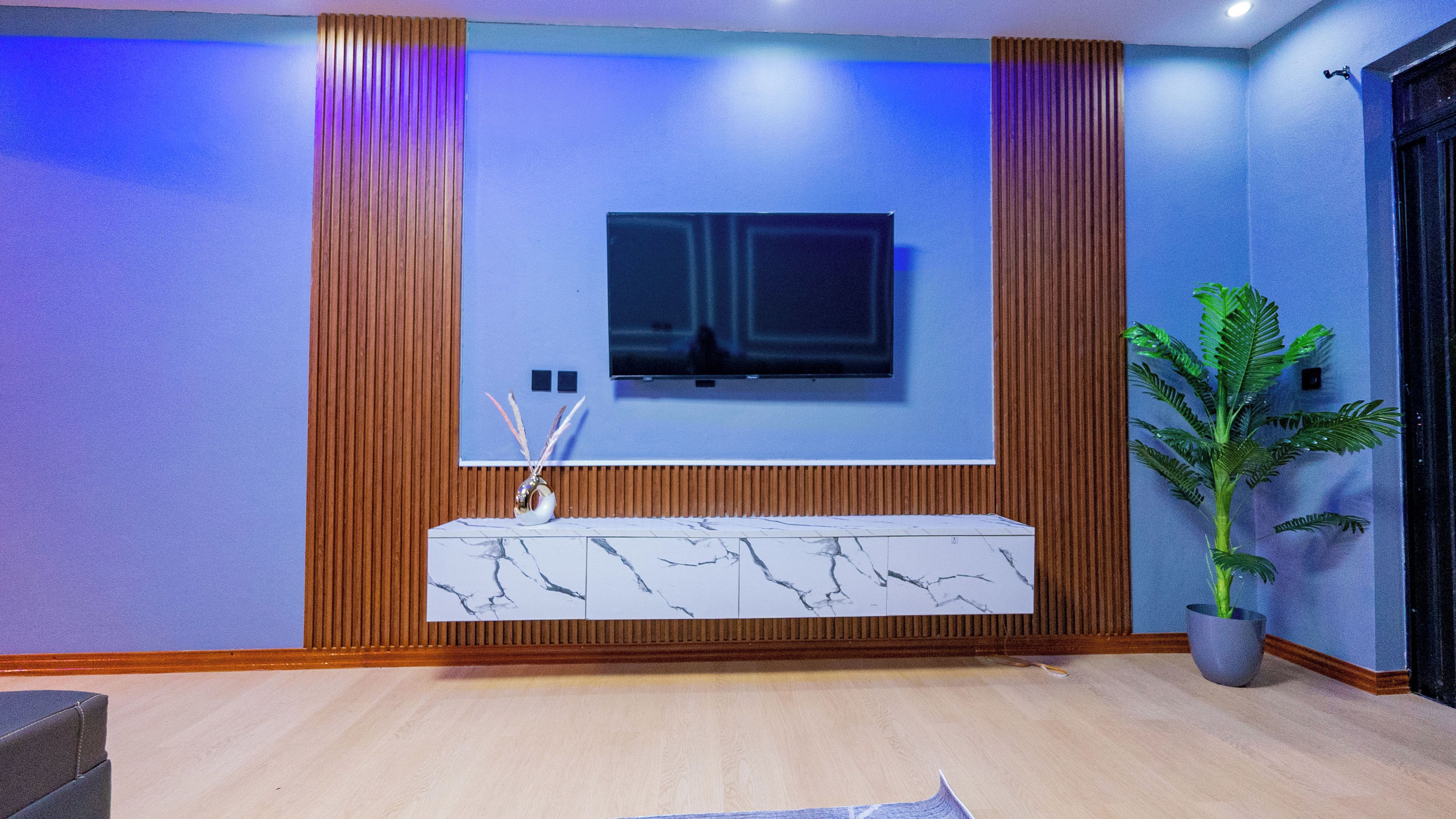Uma sala de estar moderna com uma TV de tela plana fixada em uma parede de painéis de madeira, uma mesa de console com padrão marmorizado e uma planta em vaso.