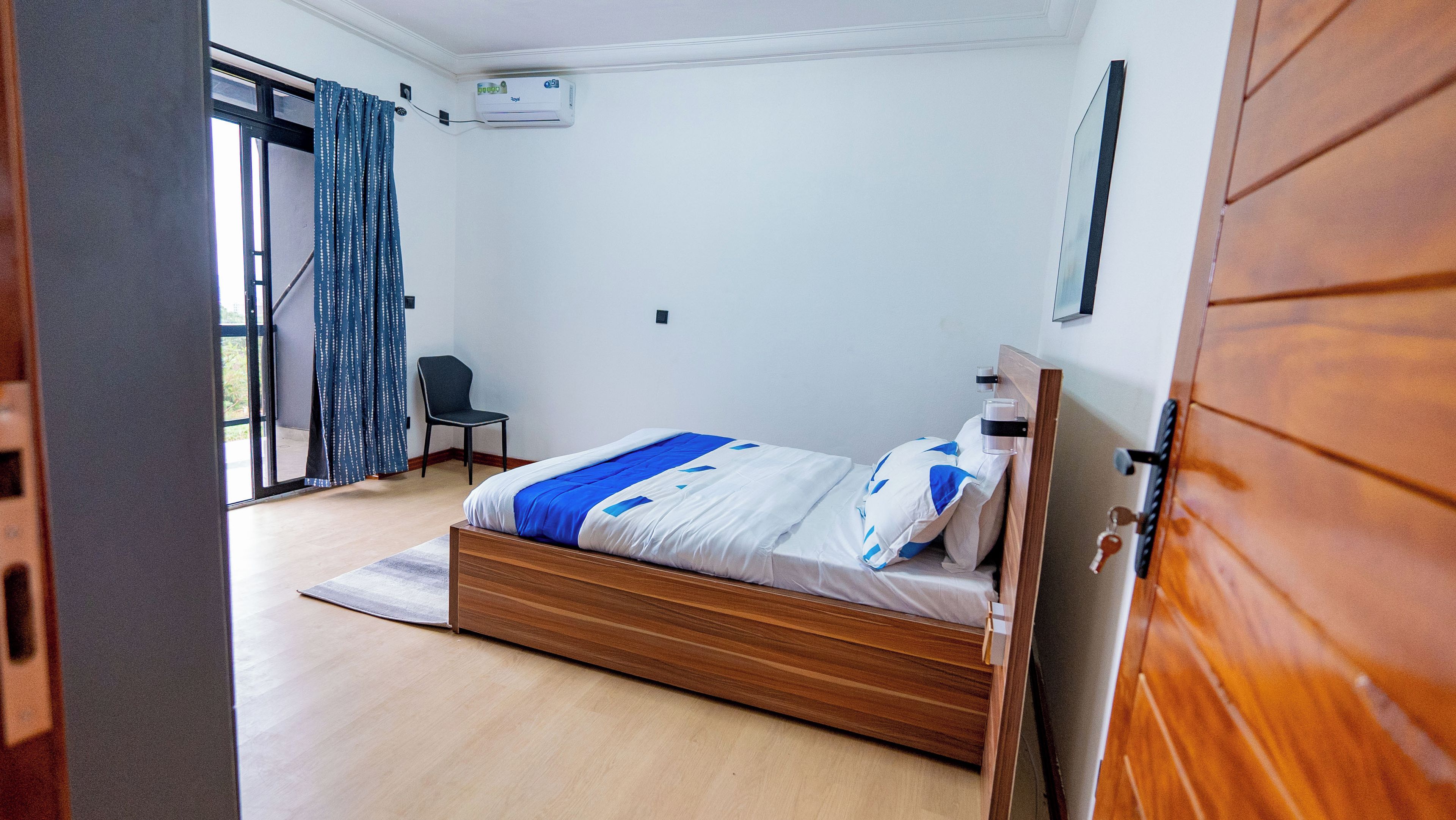 A bedroom with a wooden bed, a small chair, a window with curtains, and an air conditioning unit on the wall.