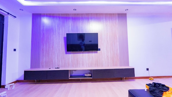 Un salon moderne doté d’un téléviseur à écran plat fixé à un mur en bois, d’un meuble de rangement intégré et d’un coin salon avec un canapé et un pouf.