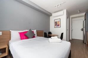 Hotellihuone, jossa on sänky, pieni pöytä, kaksi tuolia, ilmastointilaite ja ovi.