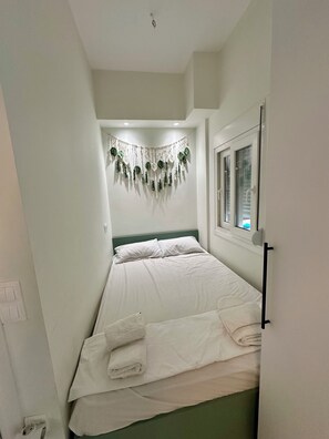 A small, neatly made bed with white linens and a headboard, a wall-mounted decorative hanging, and a window with a view.