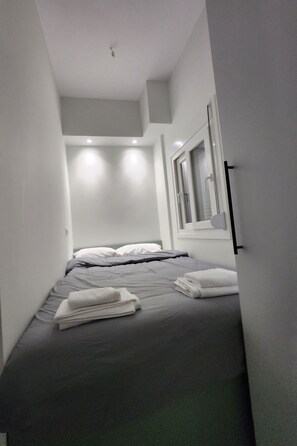 Quarto de hotel estreito com uma cama, uma janela e um espelho fixado na parede.