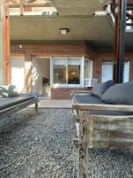 Un área de estar al aire libre con un banco de madera y un sofá, un edificio de ladrillo al fondo y un suelo de grava.