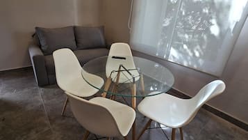 A modern living room with a glass-top dining table surrounded by four white chairs, a grey sofa with cushions, and a large window providing natural light.