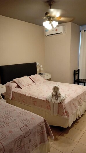 Habitación de hotel con dos camas, ventilador de techo, aire acondicionado, mesita de noche con lámpara y una silla.