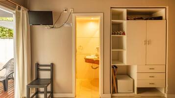 Una habitación con un televisor montado en la pared, una silla, un armario con estantes y una puerta que da a un baño.
