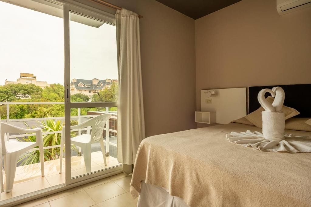 Une chambre d’hôtel avec un lit, une fenêtre avec des rideaux, une vue sur la verdure et un balcon avec des chaises.