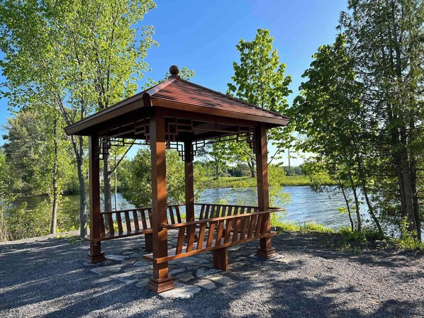 A wooden gazebo with a roof, situated by a body of water surrounded by trees.