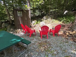 Eine Holzhütte, drei rote Stühle und ein grüner Tisch inmitten eines bewaldeten Geländes mit Steinen und Bäumen.
