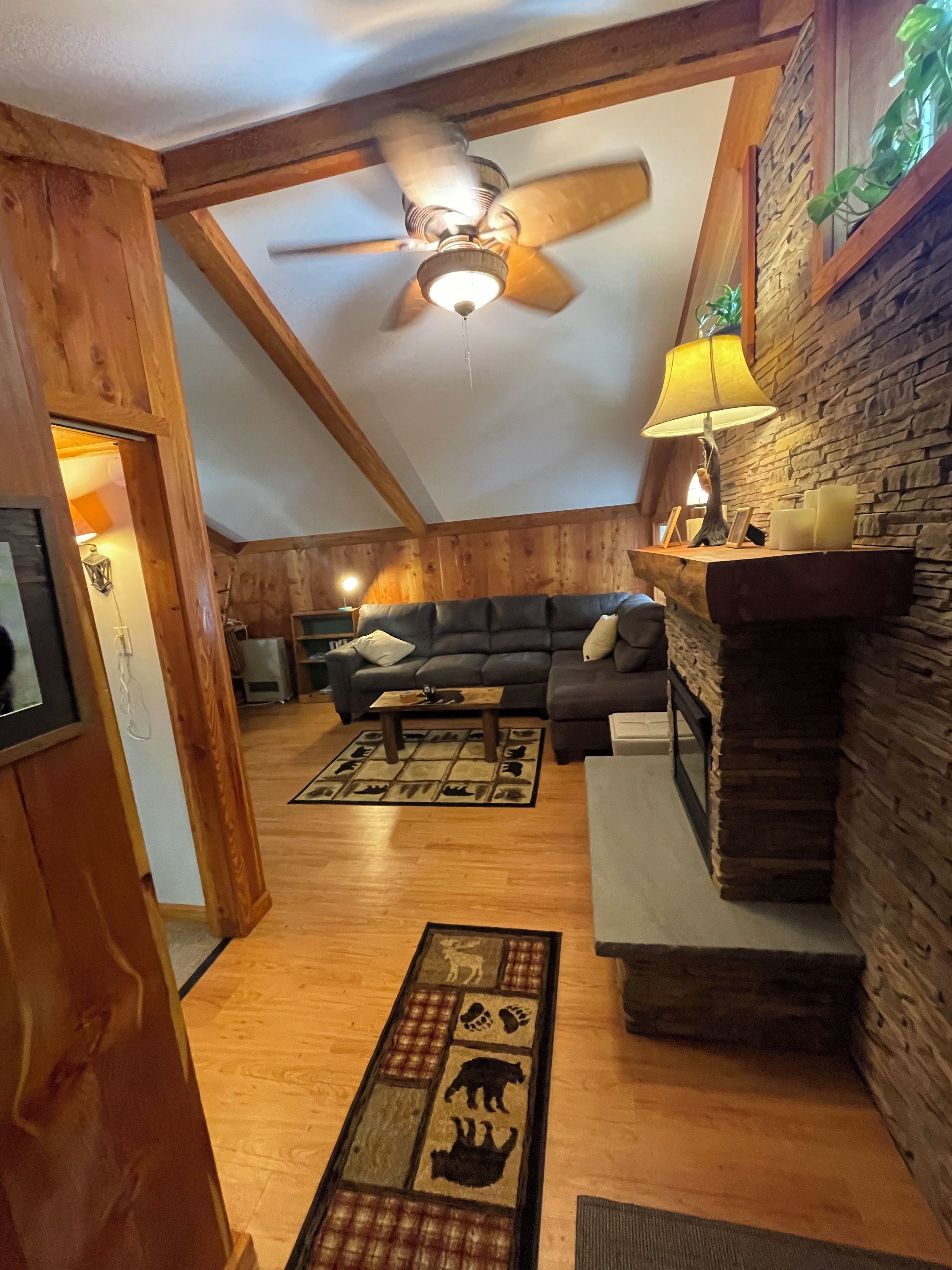 A cozy living room with a fireplace, a ceiling fan, and a stone wall.