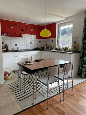 A modern kitchen with a dining area, featuring a large window, a yellow pendant light, and a red accent wall.