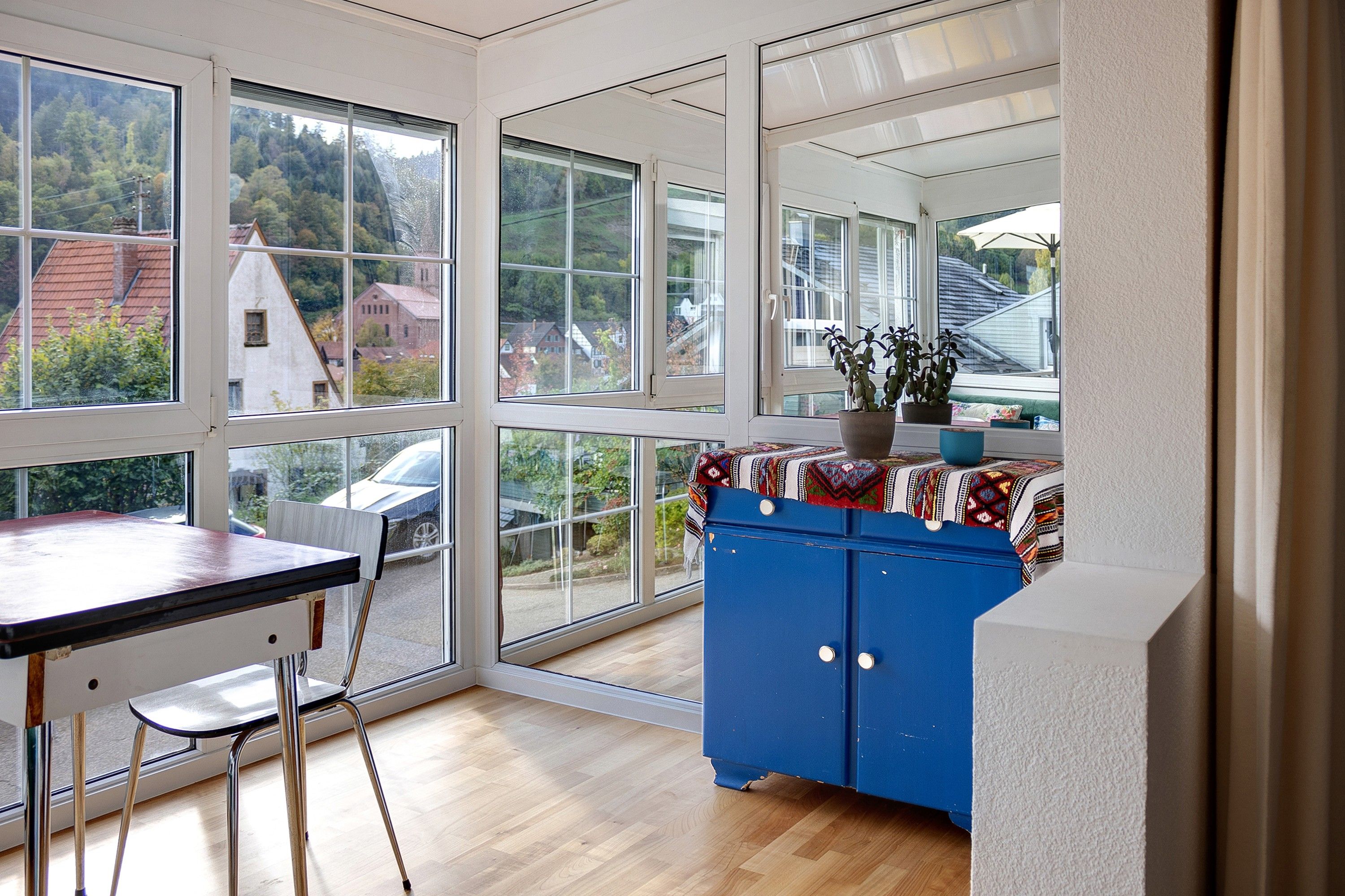 A room with large windows, a wooden table, a blue cabinet, and a view of a residential area.