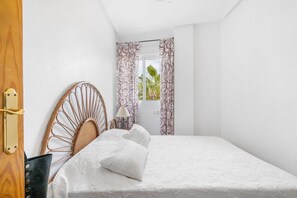 A bedroom with a bed, a wooden headboard, a nightstand with a lamp, and patterned curtains. - Tranquil Tides España  - 3 bed Appt Punta Prima (Orihuela)