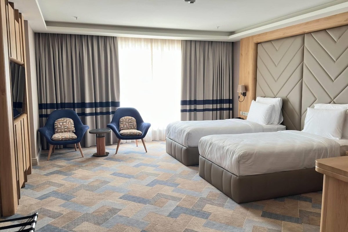 A hotel room with two beds, a seating area with chairs, a small table, and a large window with curtains.