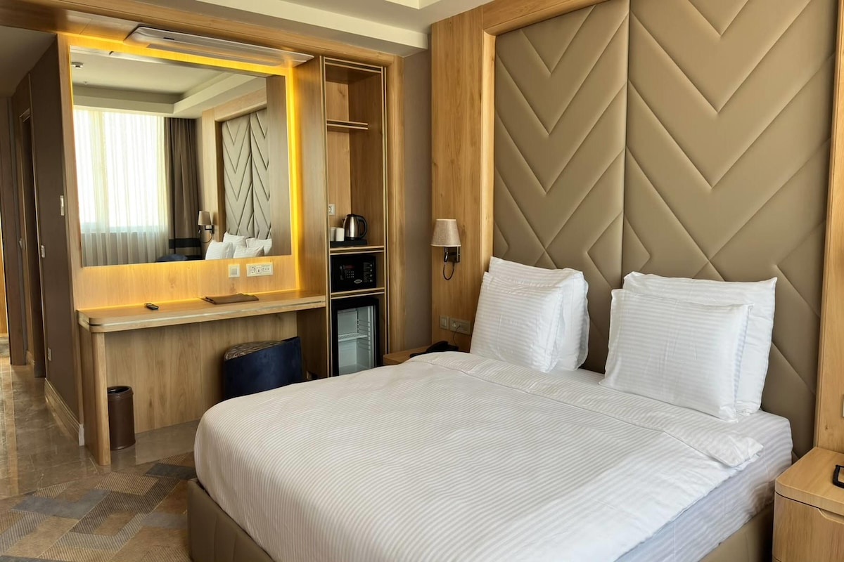 A hotel room with a bed, a desk with a mirror, a minibar, and a wall-mounted lamp.