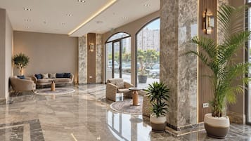 Un hall d’entrée spacieux avec un sol en marbre, un coin salon doté de canapés et de fauteuils, de grandes fenêtres offrant une vue sur la ville et des plantes en pot.