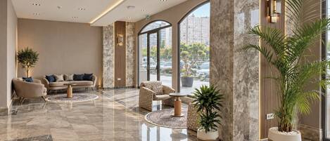 Un vestíbulo amplio con suelo de mármol, zona de estar con sofás y sillones, ventanas grandes con vistas a la ciudad y plantas en macetas.