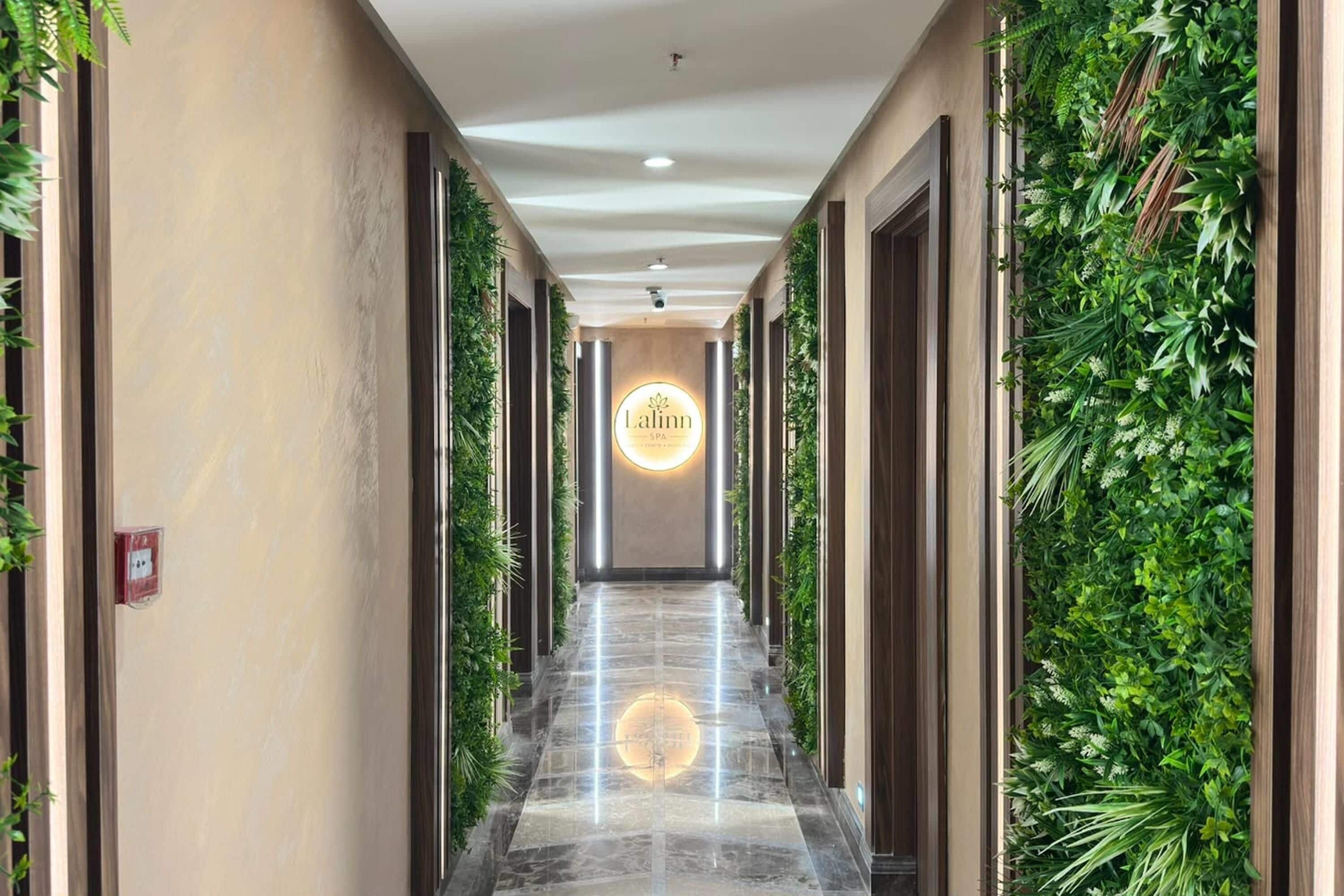 Un couloir doté d’un luminaire circulaire central, bordé de murs couverts de plantes grimpantes, qui mène à une porte sur laquelle est apposée une enseigne.
