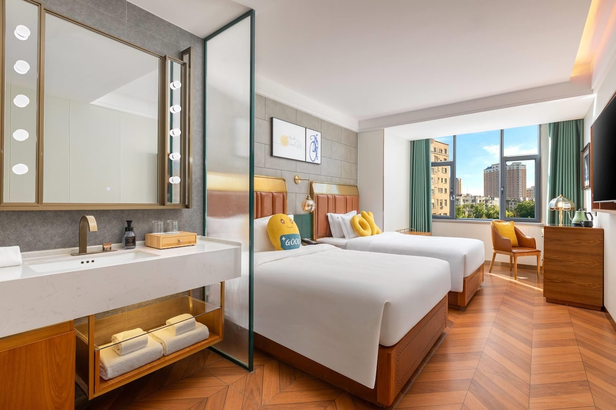A hotel room with a large mirror, two beds, a vanity with a sink, and a view of the city through large windows.