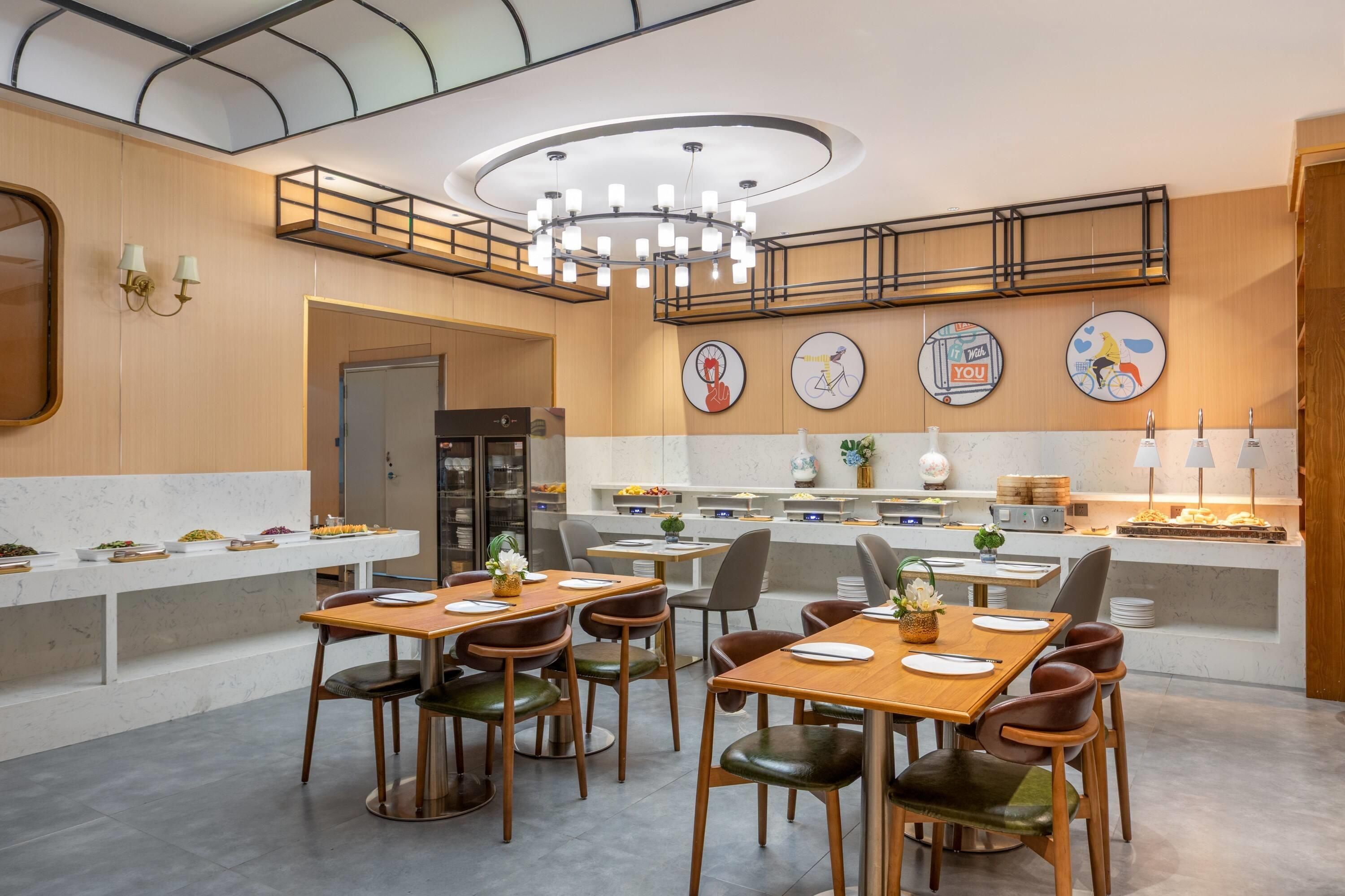Een moderne eetruimte met houten tafels en stoelen, een centraal hanglamp en een buffet met verschillende gerechten.