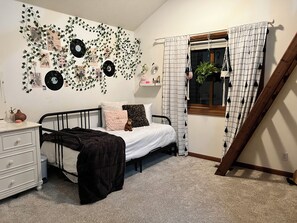 Ein Schlafzimmer mit einem Bett, einem Nachttisch, einer Kommode, einem Fenster mit Vorhängen und Wanddekorationen, darunter Schallplatten und Efeu.