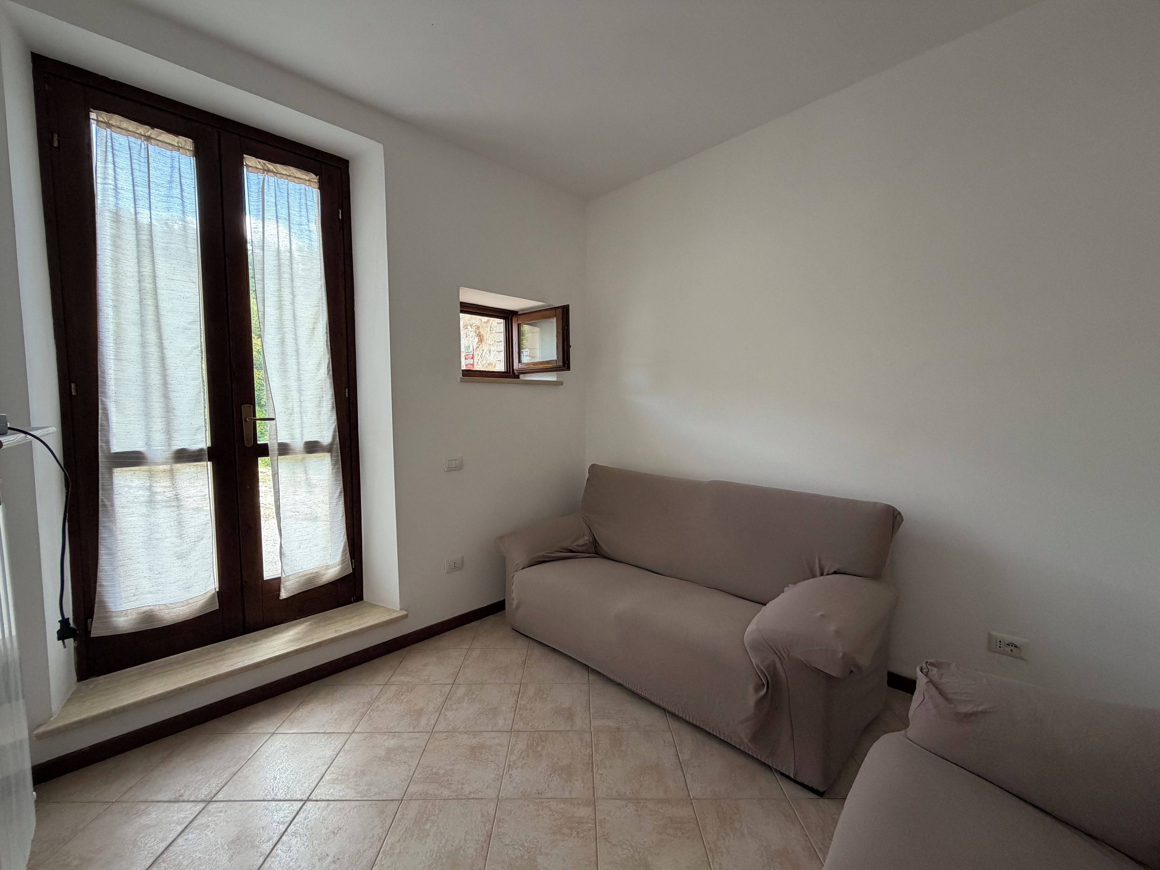 A room with a beige sofa, a small window with a mirror, and a tiled floor.