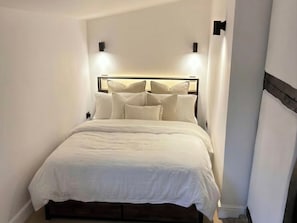 Ein ordentlich bezogenes Bett mit Kopfteil, zwei an der Wand befestigten Leuchten und einem dunkel gefärbten Nachttisch.