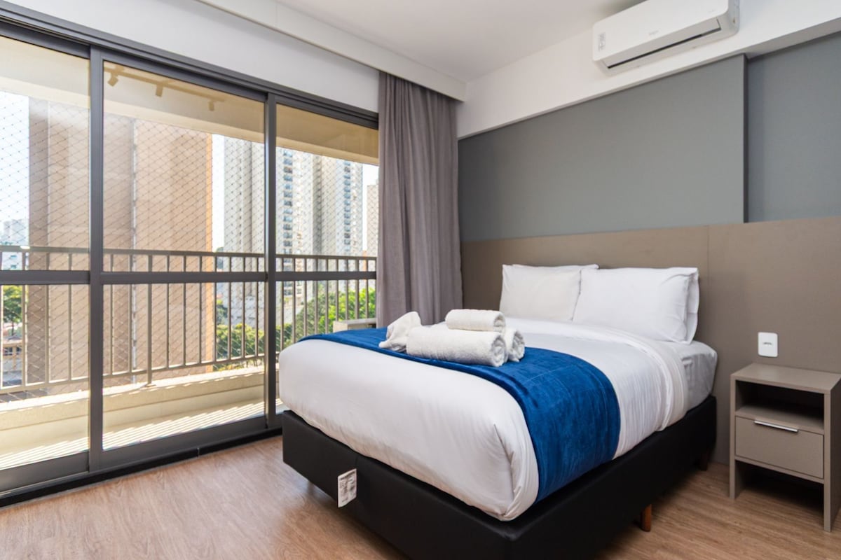 Una habitación de hotel moderna con una cama grande, una mesita de noche, aire acondicionado montado en la pared y un balcón con vistas a edificios altos.