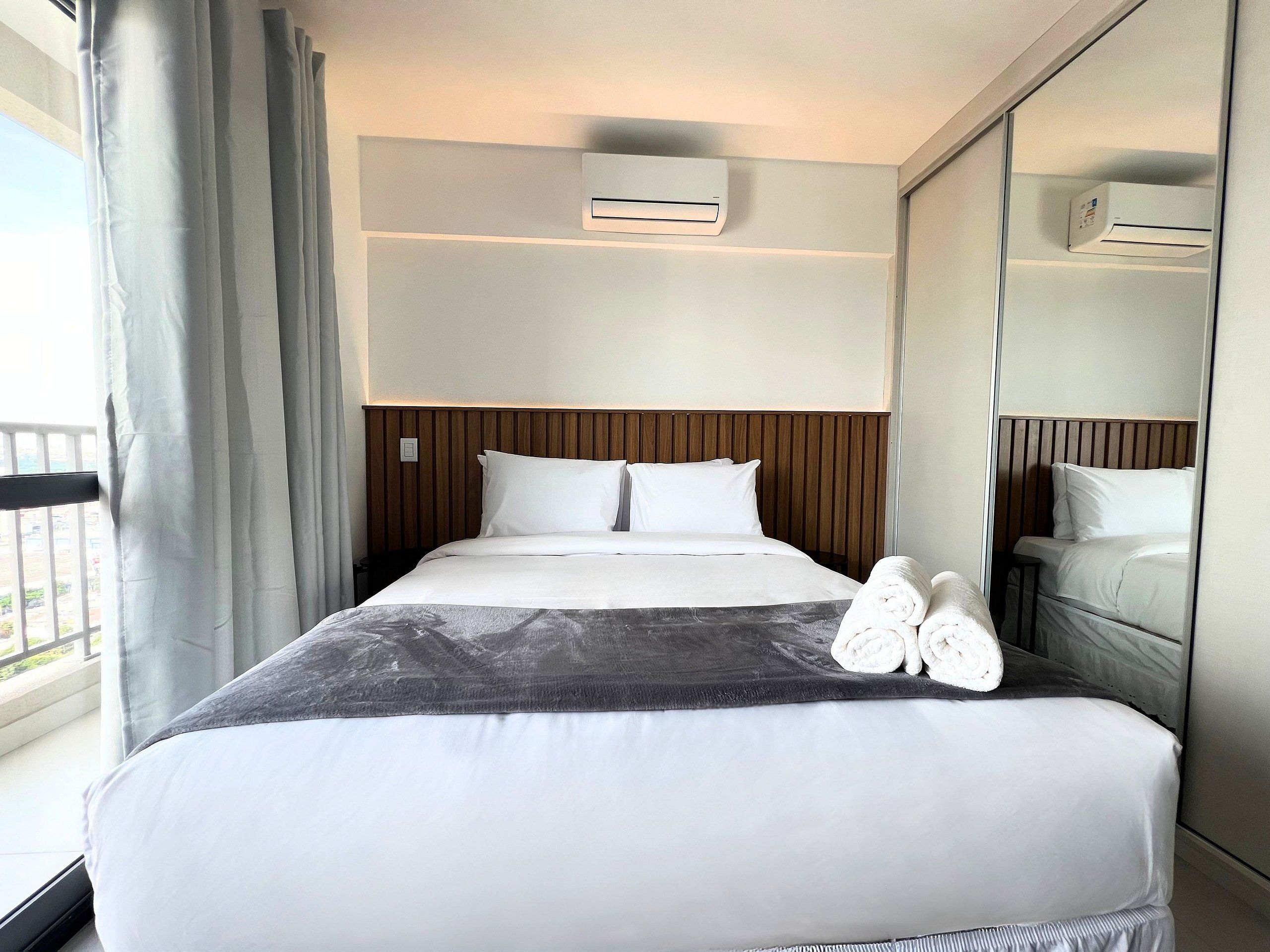 A hotel room with a bed, a bedside table, an air conditioning unit, a mirror, and a view of the outside.