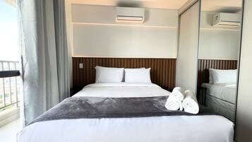 A hotel room with a bed, a bedside table, an air conditioning unit, a mirror, and a view of the outside.