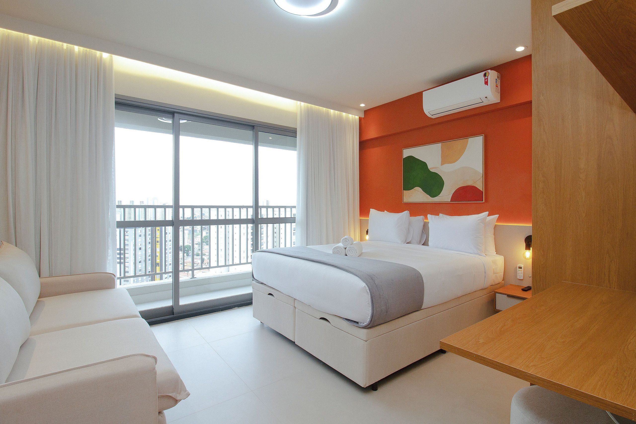 A hotel room with a bed, a desk, a chair, a bedside table, a nightstand, an air conditioning unit, and a window with a view of buildings.