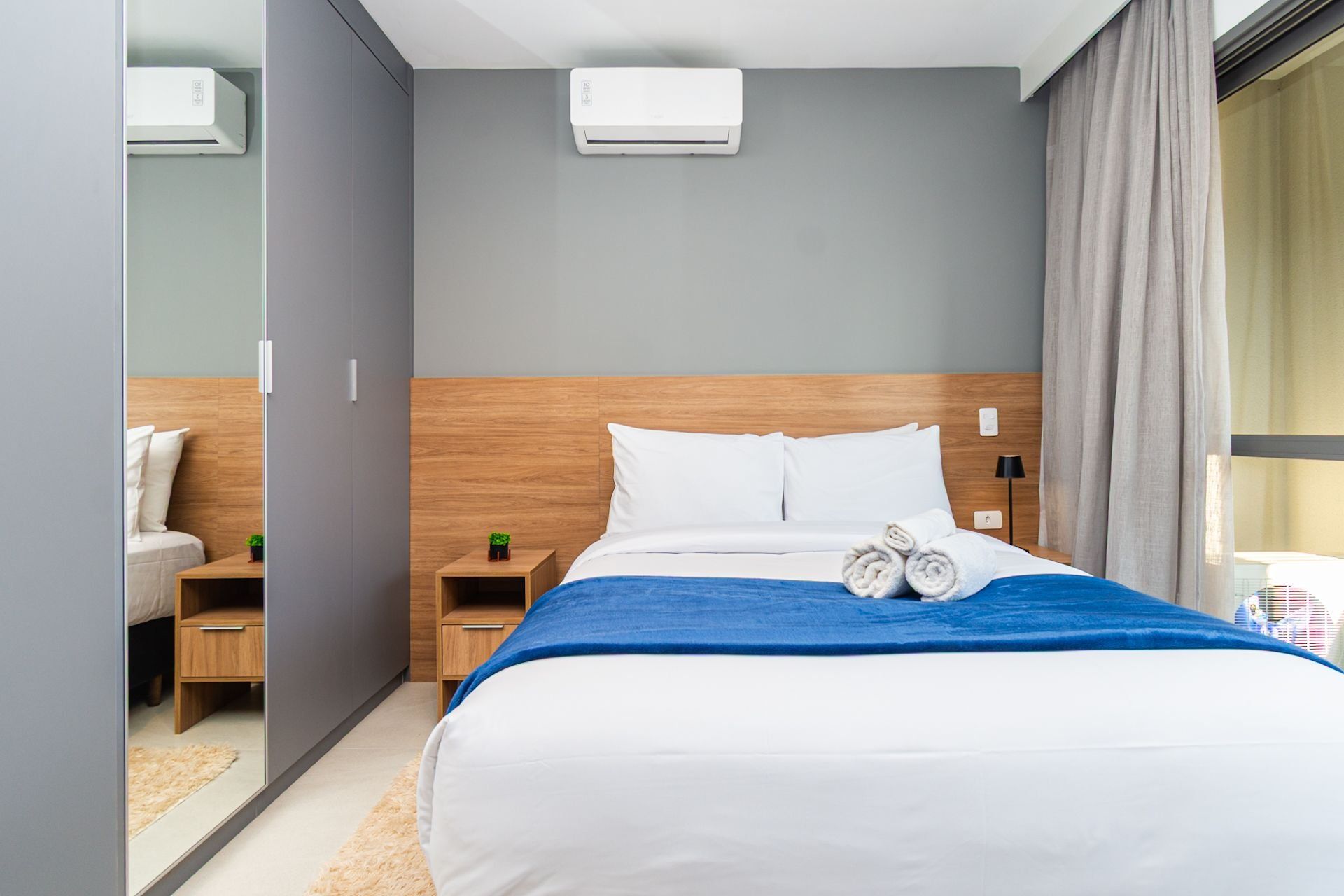 A hotel room with a bed, a nightstand, a wall-mounted air conditioner, and a window with curtains.